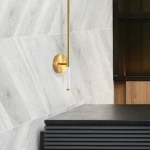 Kinkiet LED Fresco H95 o mocy 13W, barwa światła 3000K, w złotym kolorze. Posiada smukły, metalowy korpus z białym akrylowym elementem. Zainstalowany na ścianie, w nowoczesnym wnętrzu z drewnianymi i ciemnymi akcentami.