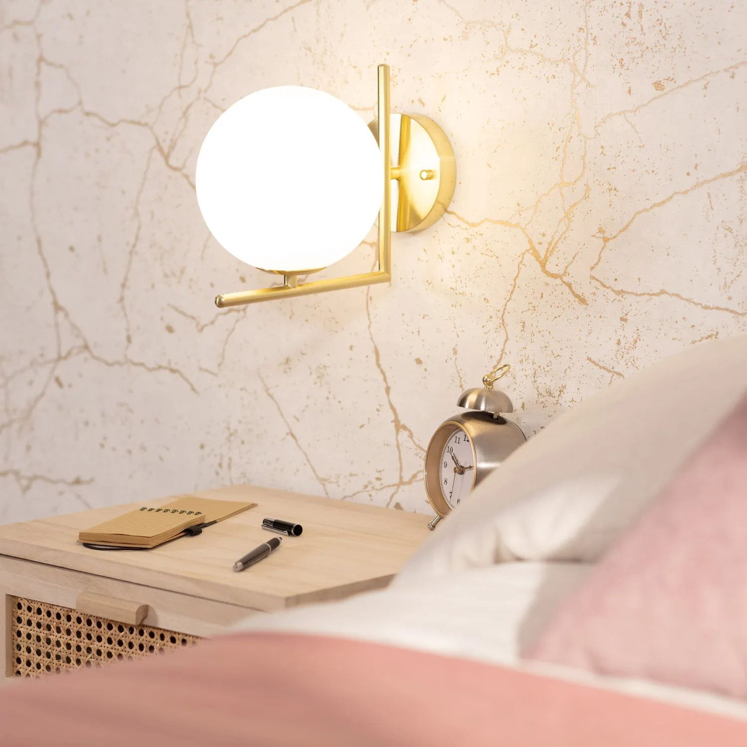 Kinkiet Riccardo-Milano 1xE14 w kolorze złotym, z okrągłym, białym kloszem. Montowany na ścianie obok stolika nocnego z drewnianą szufladą, na którym leży notes i długopisy, w tle dekoracyjna ściana z delikatnymi złotymi żyłkami.