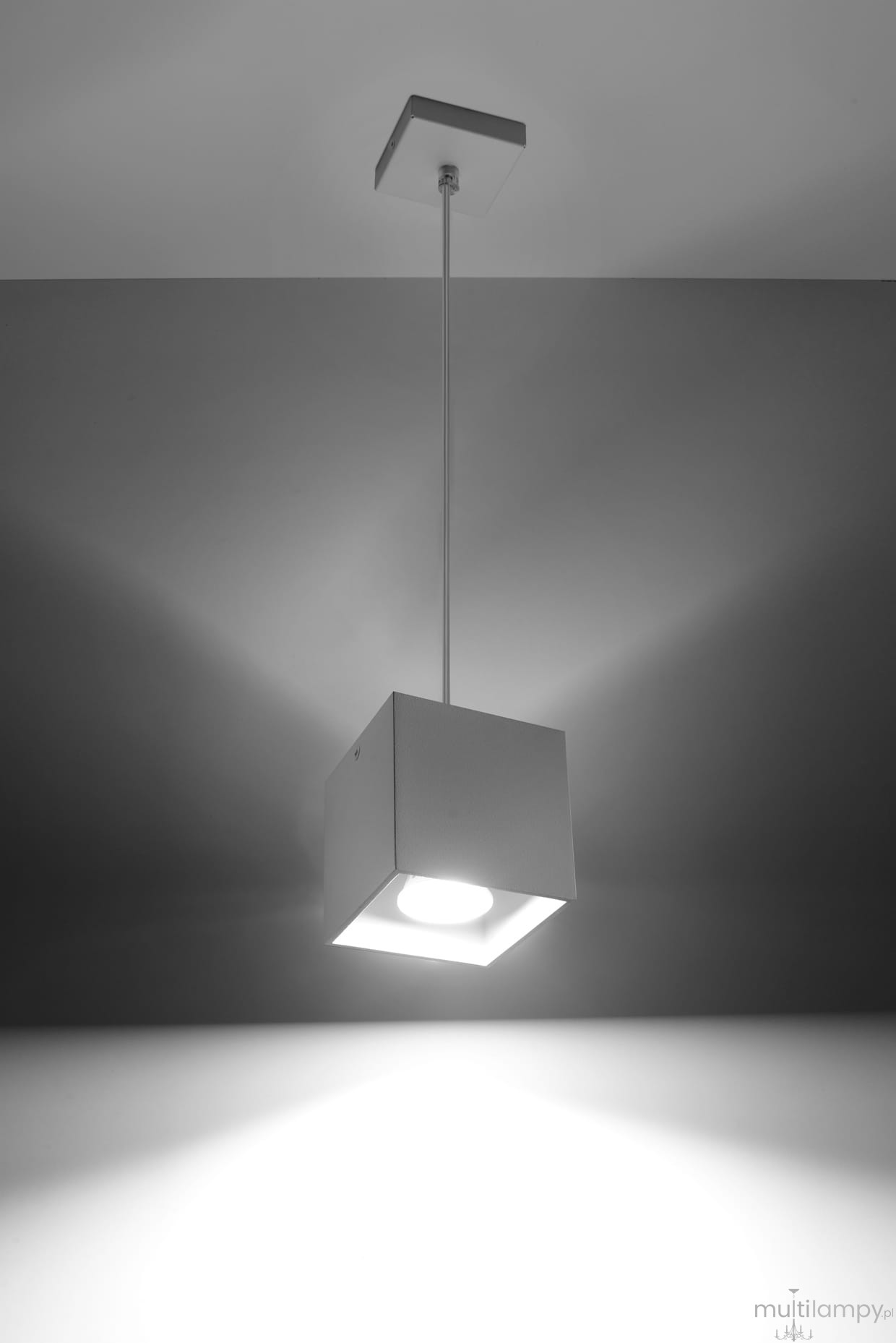 Biała, kwadratowa lampa wisząca z pojedynczym źródłem światła, zawieszona na cienkim przewodzie. Lampa ma prosty, minimalistyczny design i jest umieszczona pod kątem, co podkreśla jej nowoczesny wygląd. Tło w jasnych odcieniach.
