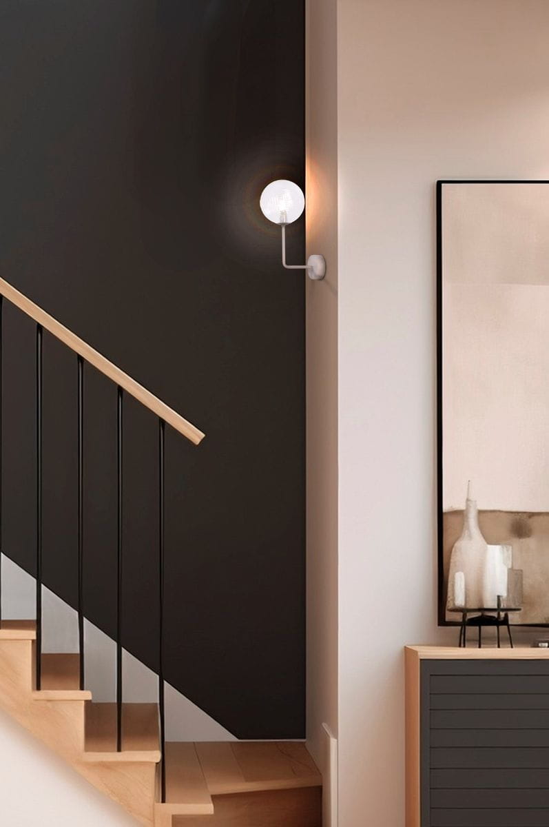 Kinkiet Casoli 1xG9 w kolorze beżowym z transparentną, okrągłą oprawą, zamontowany na ścianie w pobliżu schodów. Oświetlenie emituje ciepłe światło, a w tle widoczna jest minimalistyczna aranżacja z czarną ścianą i drewnianymi schodami.