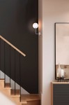 Kinkiet Casoli 1xG9 w kolorze beżowym z transparentną, okrągłą oprawą, zamontowany na ścianie w pobliżu schodów. Oświetlenie emituje ciepłe światło, a w tle widoczna jest minimalistyczna aranżacja z czarną ścianą i drewnianymi schodami.