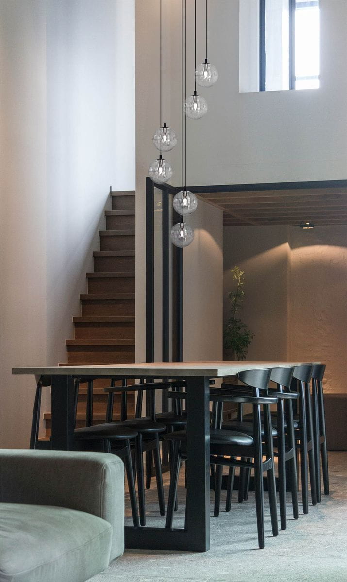 Lampa wisząca Monza w kolorze czarnym i grafitowym, z sześcioma kloszami G9, zwisająca nad stołem w nowoczesnej jadalni. Widoczna w aranżacji z drewnianymi schodami i czarnymi krzesłami wokół stołu.