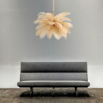 Lampa wisząca Emu w kolorze złotym i beżowym, z 6 miejscami na żarówki E14, o formie przypominającej pióra. Zawieszona nad szarym sofą, w minimalistycznym wnętrzu z gładką ścianą. Wykonana z lekkiego materiału, dekoracyjna i nowoczesna.