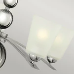 Lampa wisząca Zelda w kolorze polerowanego niklu z pięcioma kloszami z matowego szkła, umieszczona na metalowej konstrukcji. Widoczne zaokrąglone elementy i eleganckie detale. Klosze skierowane w dół, zamontowane na łukowatych ramionach.