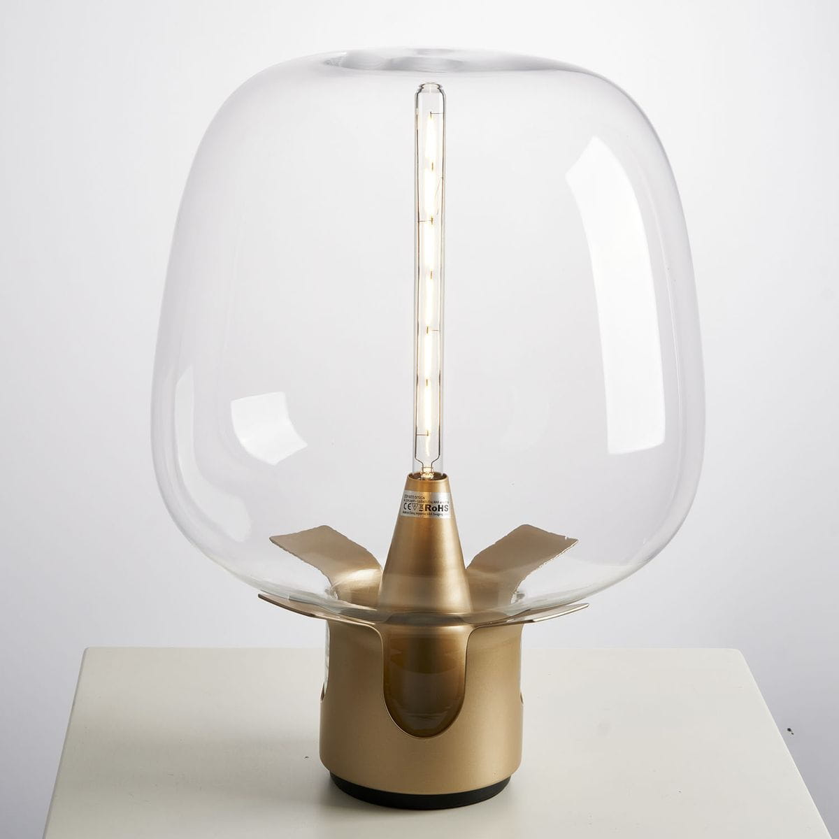 Lampa stołowa Block 1xE27 w złotym kolorze, z przezroczystym kloszem i metalową podstawą. Ujęcie frontalne, lampka stoi na białym stole, w środku widoczna żarówka w kształcie rury.