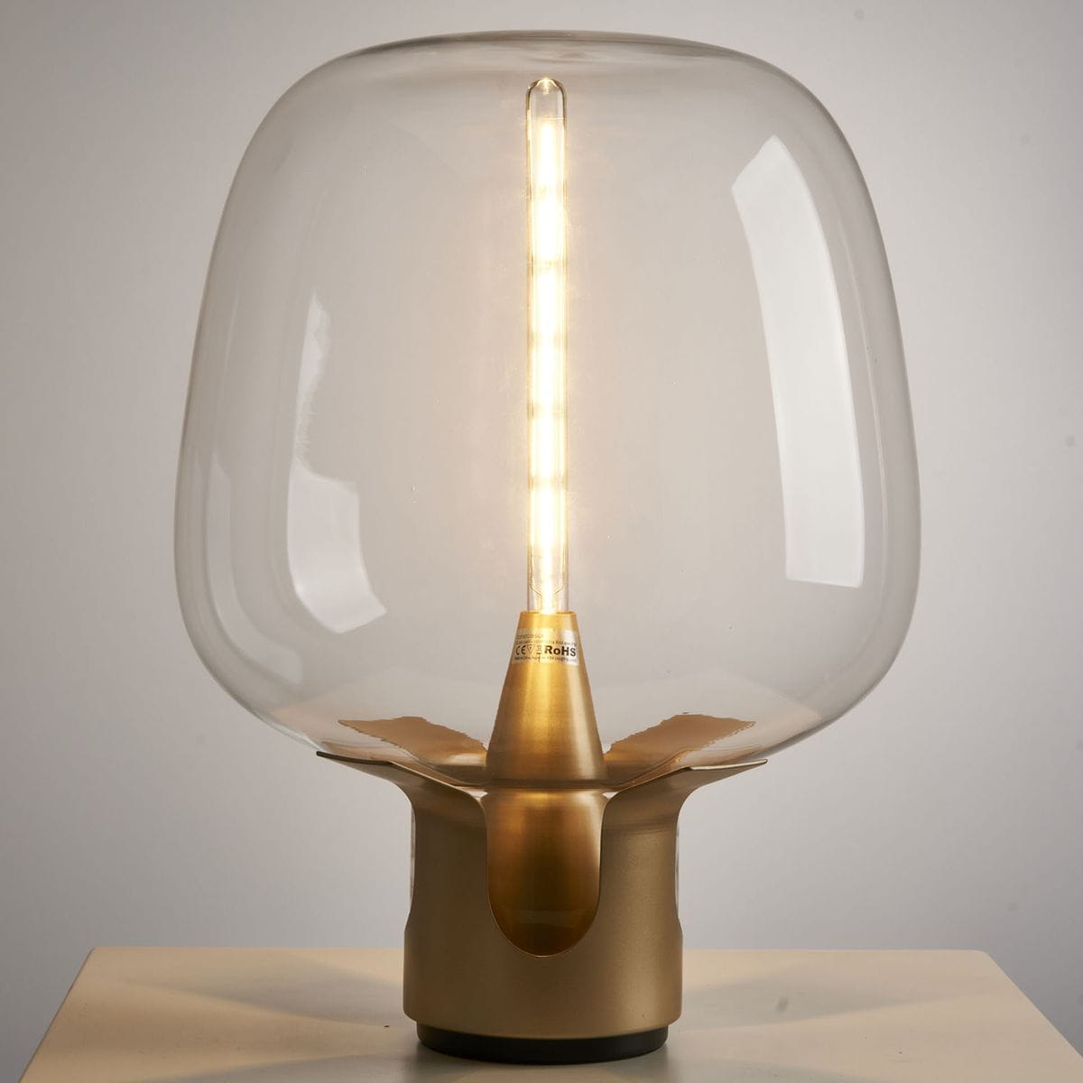 Lampa stołowa Block z żarówką E27, z przezroczystym kloszem w kształcie gruszki i złotą podstawą. W centralnej części widoczna diodowa żarówka, emitująca ciepłe światło. Produkt ustawiony na białym tle, w pozycji stojącej.