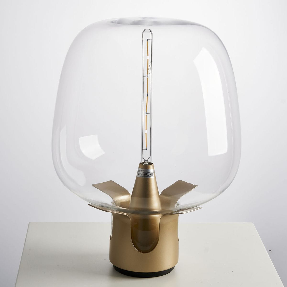 Lampa stołowa Block 1xE27 w kolorze złotym z przezroczystym kloszem. Ujęcie frontalne, lampa stoi na płaskiej powierzchni. Widoczna żarówka w środku oraz elegancka, złota podstawa z detalami. Tło białe, proste.