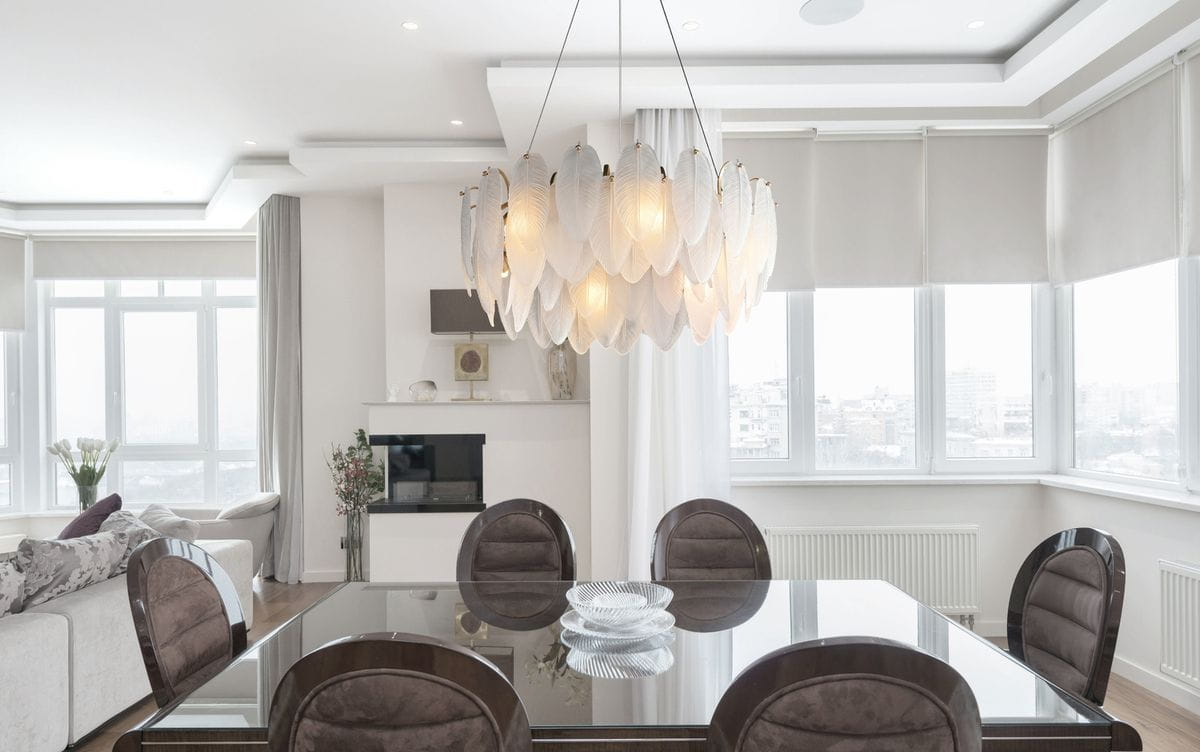 Lampa wisząca Piuma w kolorze mosiądzu z 10 gniazdami E14. Ma oprawę w kształcie liści, zawieszoną na cienkim kablu. Widoczna w nowoczesnym wnętrzu, nad okrągłym stołem, w otoczeniu krzeseł i dekoracyjnych elementów.