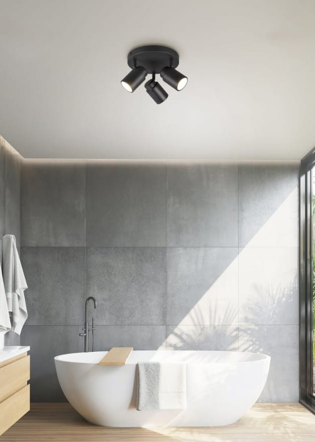 Czarna lampa sufitowa Angelo IP44 z trzema reflektorami GU10, zamontowana na suficie. Reflektory skierowane w różnych kierunkach. Lampa w nowoczesnej łazience z jasnym tłem, wanną i drewnianą podłogą.