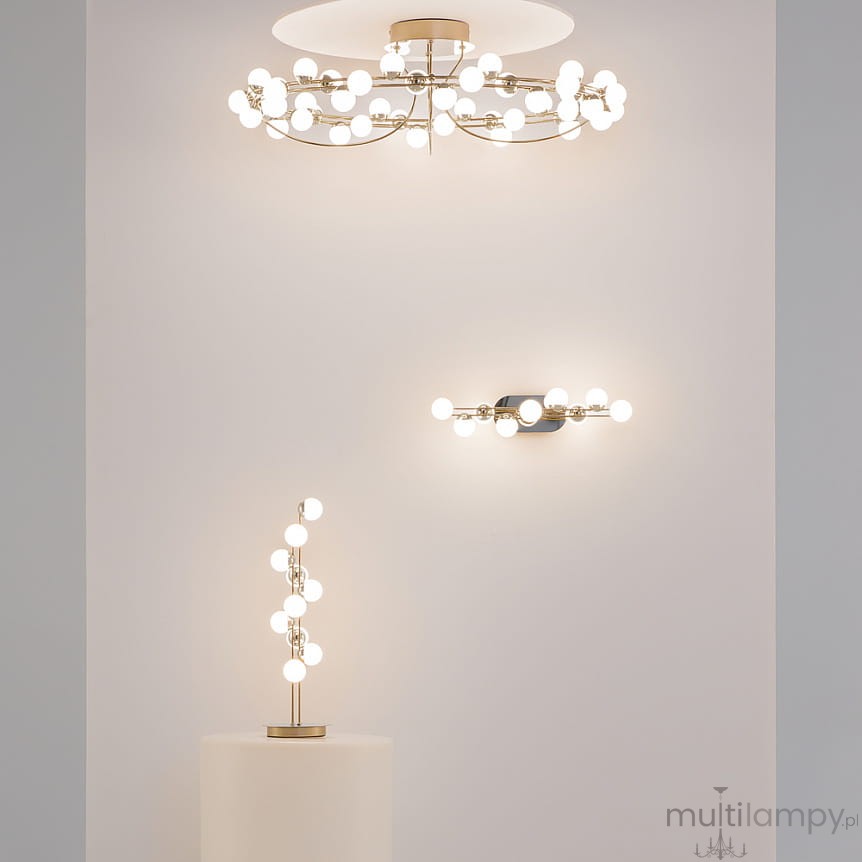 Złota lampa sufitowa LED Tessara o mocy 72W i temperaturze barwowej 3000K, z okrągłym kloszem i dekoracyjnymi elementami, zawieszona na ścianie. W tle widoczna jest lampa stojąca oraz kinkiet w podobnym stylu.