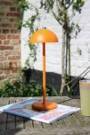 Pomarańczowa lampa stołowa Joy LED, o zaokrąglonym kloszu i smukłym stelażu, stoi na drewnianym stole obok magazynu i filiżanki. W tle widoczna jest zieleń roślin oraz ceglana ściana, co tworzy przyjemny, zewnętrzny kontekst.