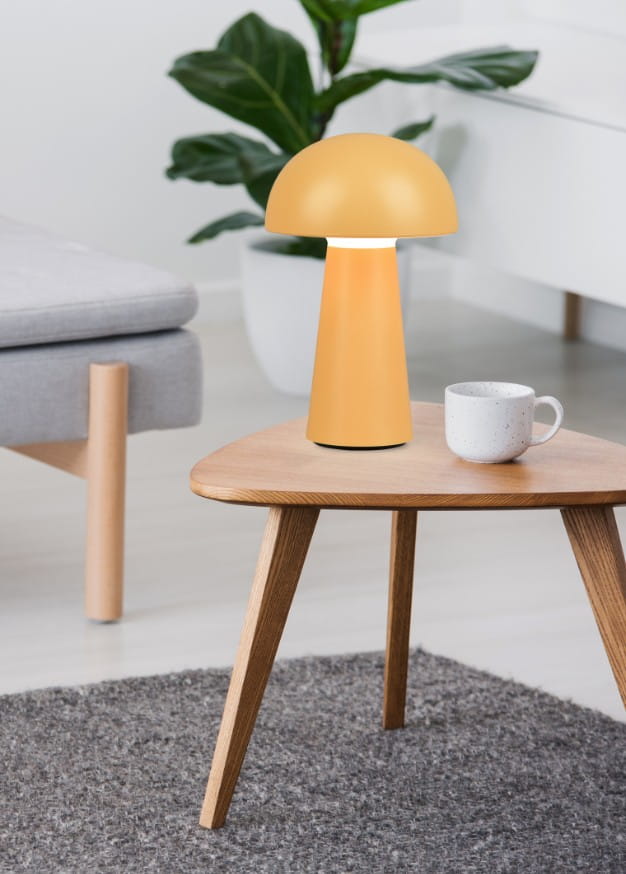 Pomarańczowa lampa stołowa Lennon LED o kształcie grzyba, wykonana z matowego materiału. Ustawiona na drewnianym stoliku, obok białej filiżanki. Tło z rośliną i szarym dywanem tworzy przytulną atmosferę.
