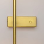 Mosiężny kinkiet LED z dotykowym włącznikiem, wykończony w matowym złocie. Widoczny prosty kształt z długą, smukłą nogą oraz płaską podstawą z symbolem włącznika. Produkt zamontowany na jasnym tle.