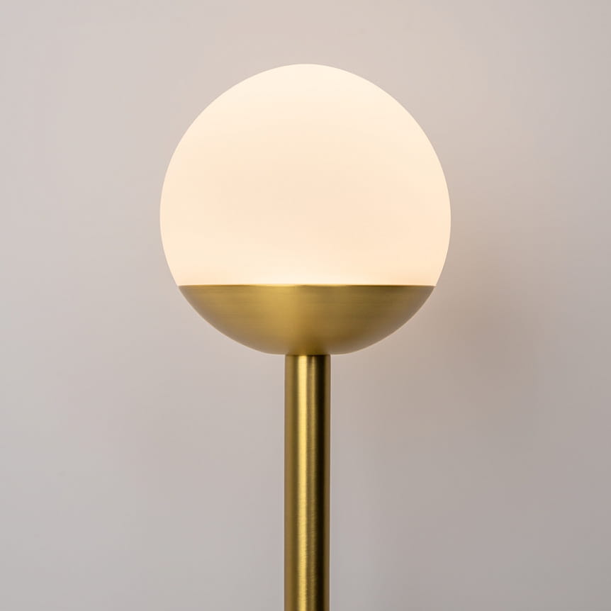 Kinkiet LED w kolorze mosiądzu z okrągłą, białą kloszem, umieszczony na prostym, cylindrycznym stelażu. Klosz ma matowe wykończenie, a lampa emituje ciepłe światło (3000K). Ujęcie frontalne, na jasnym tle.