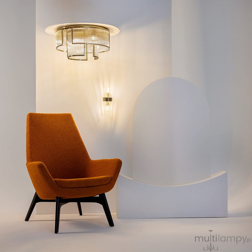 Lampa sufitowa Frame 9xE14 w kolorze złotym, o geometrycznym kształcie, zawieszona na okrągłej podstawie. Widoczne są również detale szklanych kloszy i stylowy design. Lampa umieszczona w nowoczesnym wnętrzu z pomarańczowym fotelem.