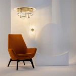 Lampa sufitowa Frame 9xE14 w kolorze złotym, o geometrycznym kształcie, zawieszona na okrągłej podstawie. Widoczne są również detale szklanych kloszy i stylowy design. Lampa umieszczona w nowoczesnym wnętrzu z pomarańczowym fotelem.