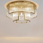 Lampa sufitowa Frame z dziewięcioma gniazdami E14 w złotej oprawie. Okrągły klosz z przezroczystego materiału z pionowymi żłobieniami, zawieszony pod sufitem, emitujący jasne światło. Prosta, elegancka forma w nowoczesnym stylu.