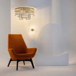 Lampa sufitowa Frame w kolorze złotym, z sześcioma miejscami na żarówki E14, o nowoczesnym, geometrycznym kształcie i metalowej konstrukcji. Zawieszona na białym tle, w otoczeniu pomarańczowego fotela i subtelnych detali architektonicznych.