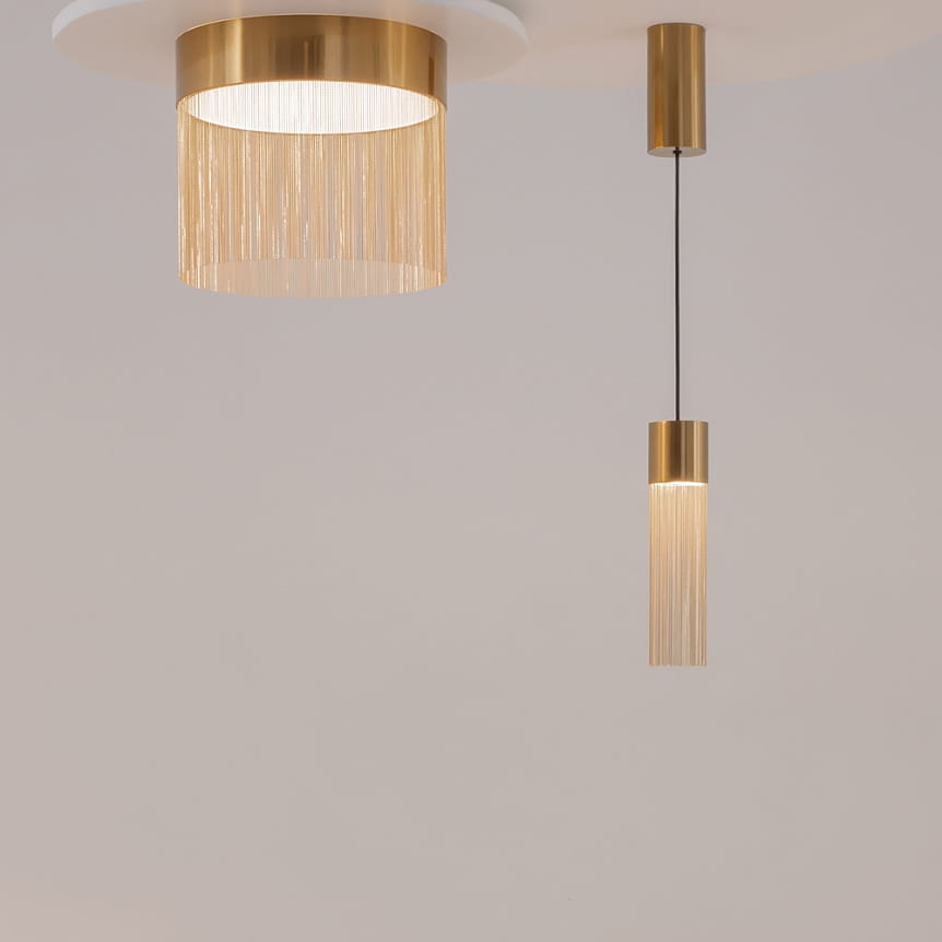 Lampa wisząca LED Imaginary w kolorze złotym, z głównym kloszem w formie cylindrycznej z frędzlami oraz mniejszym, prostym kloszem. Oświetlenie ma moc 5W i temperaturę barwową 3000K, zawieszona na cienkim przewodzie, z jasnym tłem.