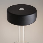 Lampa stołowa LED AI Collaboration MOD229TL-L3B3K1 z czarnym, matowym kloszem w kształcie dysku i przezroczystą podstawą. Ujęcie z góry, ukazujące centralny włącznik. Tło w neutralnym kolorze.