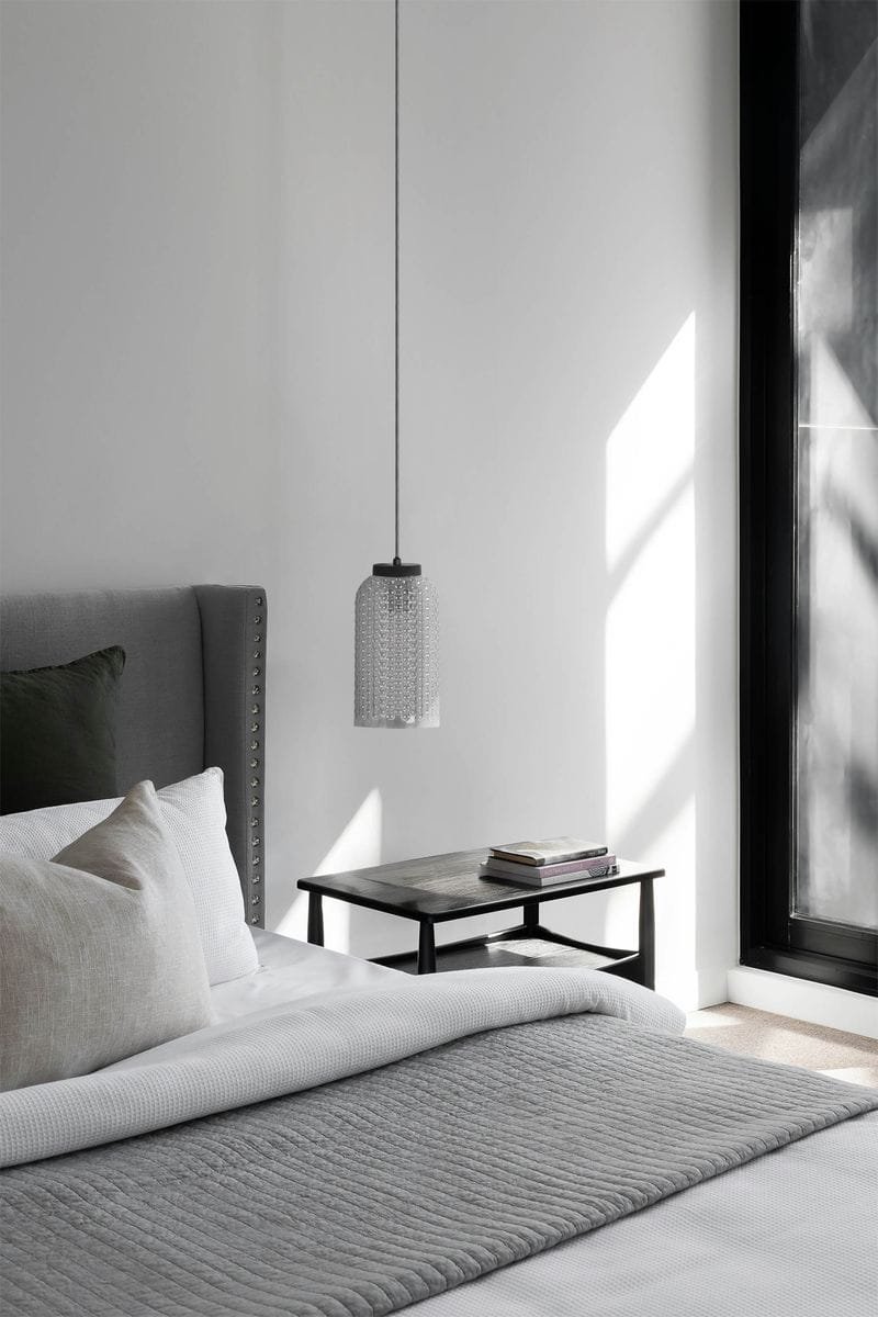 Lampa wisząca Inga D14 z kloszem w kolorze grafitowym, na czarnym przewodzie. Zawieszona nad drewnianym stolikiem w jasnym wnętrzu, obok szarego łóżka z poduszkami. Klosz ma teksturowaną powierzchnię, co dodaje elegancji.