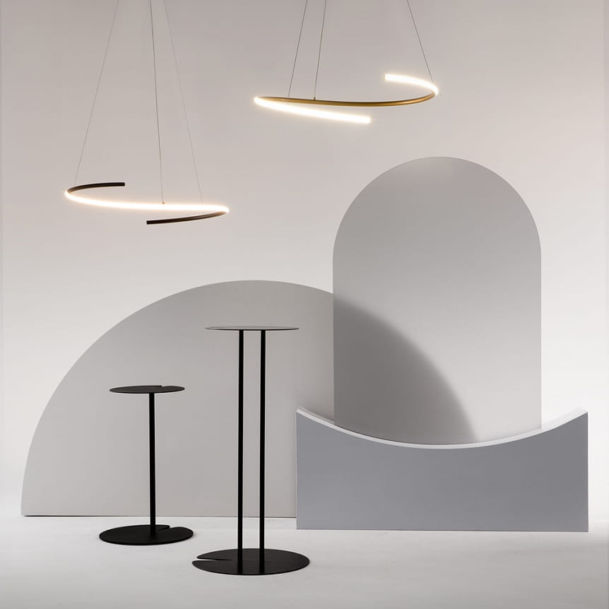 Lampa wisząca LED Curve w kolorze mosiądzu, o mocy 31W, zawieszona w nowoczesnym wnętrzu. Dwa okrągłe źródła światła o minimalistycznym kształcie na tle geometrycznych form w odcieniach szarości. Wysokość lampy regulowana.