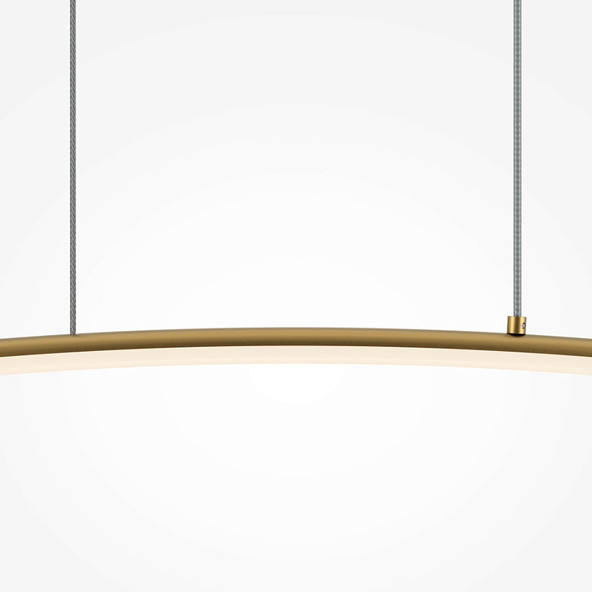 Lampa wisząca LED Curve w kolorze mosiężnym, o prostym, zakrzywionym kształcie. Zawieszona na dwóch cienkich przewodach, z delikatnym, białym źródłem światła w dolnej części. Tło neutralne, jasne.