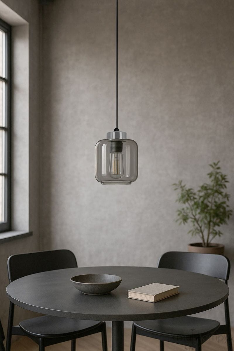 Lampa wisząca Piega 1xE27 w kolorze czarnym i grafitowym, w kształcie prostopadłościanu z przezroczystym kloszem. Zawieszona na długim, czarnym przewodzie nad okrągłym stołem, obok ceramicznej miski i książki, w minimalistycznym wnętrzu.