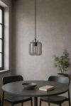 Lampa wisząca Piega 1xE27 w kolorze czarnym i grafitowym, w kształcie prostopadłościanu z przezroczystym kloszem. Zawieszona na długim, czarnym przewodzie nad okrągłym stołem, obok ceramicznej miski i książki, w minimalistycznym wnętrzu.