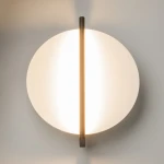 Kinkiet LED Mira, moc 14W, kolor czarny. Okrągła, matowa obudowa z białego materiału oraz centralna, pionowa linia emitująca ciepłe światło (3000K). Zawieszony na ścianie, minimalistyczny design.