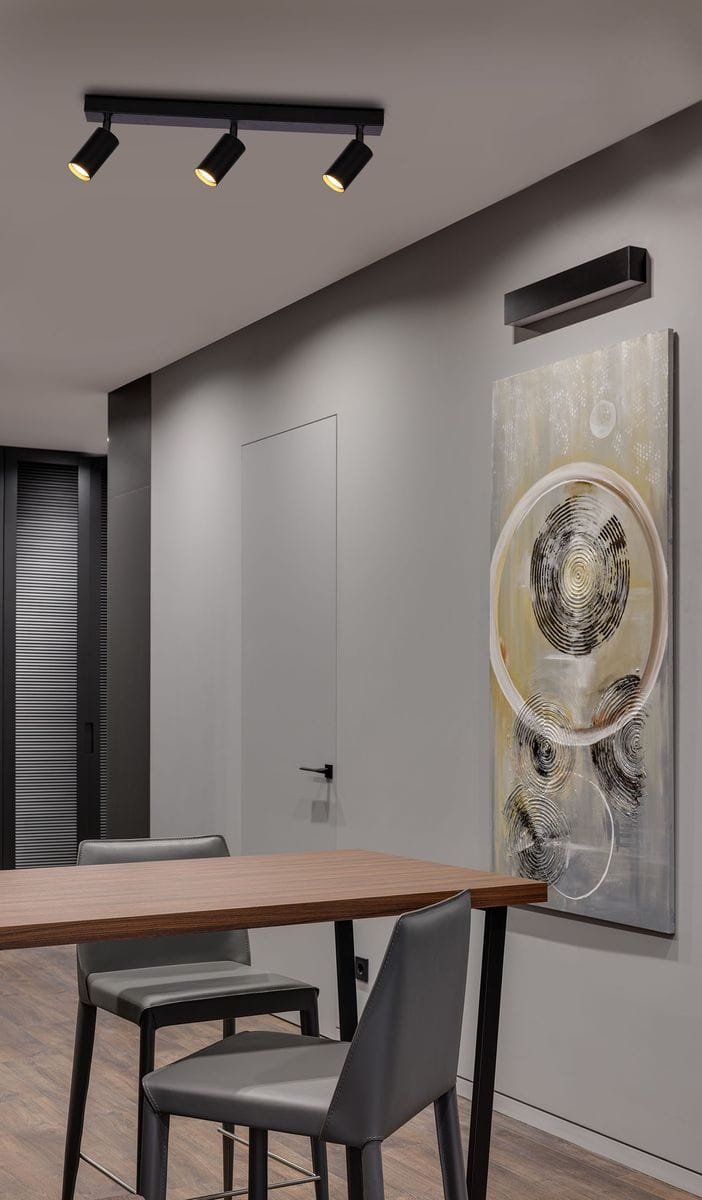 Monti lampa sufitowa w kolorze czarnym z trzema reflektorami GU10. Ujęcie z góry, zamontowana na suficie nad drewnianym stołem i krzesłami, w nowoczesnym wnętrzu z dekoracyjnym obrazem na ścianie.
