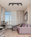 Czarna lampa sufitowa Monti z czterema spotami GU10, zamontowana równolegle do sufitu. Lampa znajduje się w jasnym pomieszczeniu z dużymi oknami, obok różowego sofy i szklanej ławy. W tle firany i dekoracyjne obrazy.