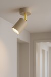 Kinkiet Mati w kolorze beżowym z jedną oprawą GU10, zamontowany na suficie. Lampa w kształcie cylindrycznym, z metalowym wykończeniem w odcieniu złotym, skierowana w dół. Tło jasne, minimalistyczne wnętrze.