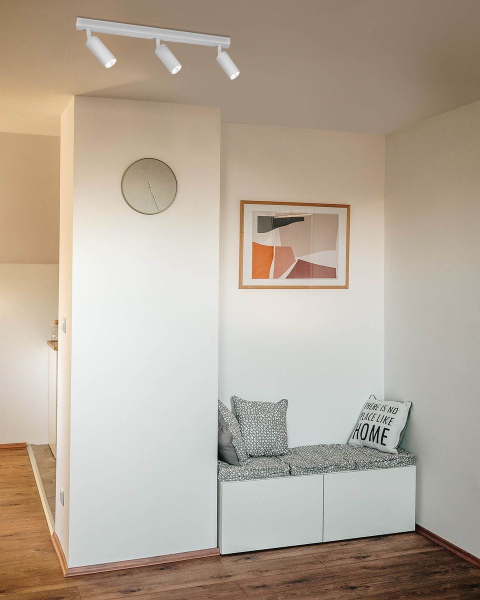 Biała lampa sufitowa Kayla z trzema reflektorami GU10, zamontowana na ścianie w narożniku pokoju. Lampa o prostym, nowoczesnym designie, oświetlająca przestrzeń nad miejscem do siedzenia z poduszkami.