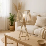Beżowa lampa stołowa Livia z metalowym kloszem w formie siatki, z żarówką E27 w środku. Ustawiona na drewnianym stole, w tle widoczna kanapa z poduszkami i rośliny dekoracyjne.