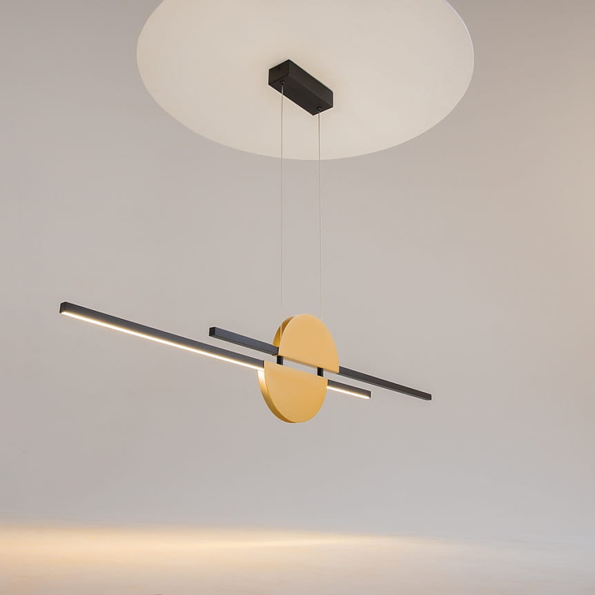 Lampa wisząca Skyline LED o mocy 26W, z okrągłym elementem w kolorze złotym oraz czarnymi, prostokątnymi ramionami. Zawieszona na cienkich linkach, z okrągłym, białym podsufitką. Wnętrze minimalistyczne, tło jasne.