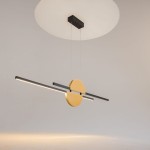Lampa wisząca Skyline LED o mocy 26W, z okrągłym elementem w kolorze złotym oraz czarnymi, prostokątnymi ramionami. Zawieszona na cienkich linkach, z okrągłym, białym podsufitką. Wnętrze minimalistyczne, tło jasne.