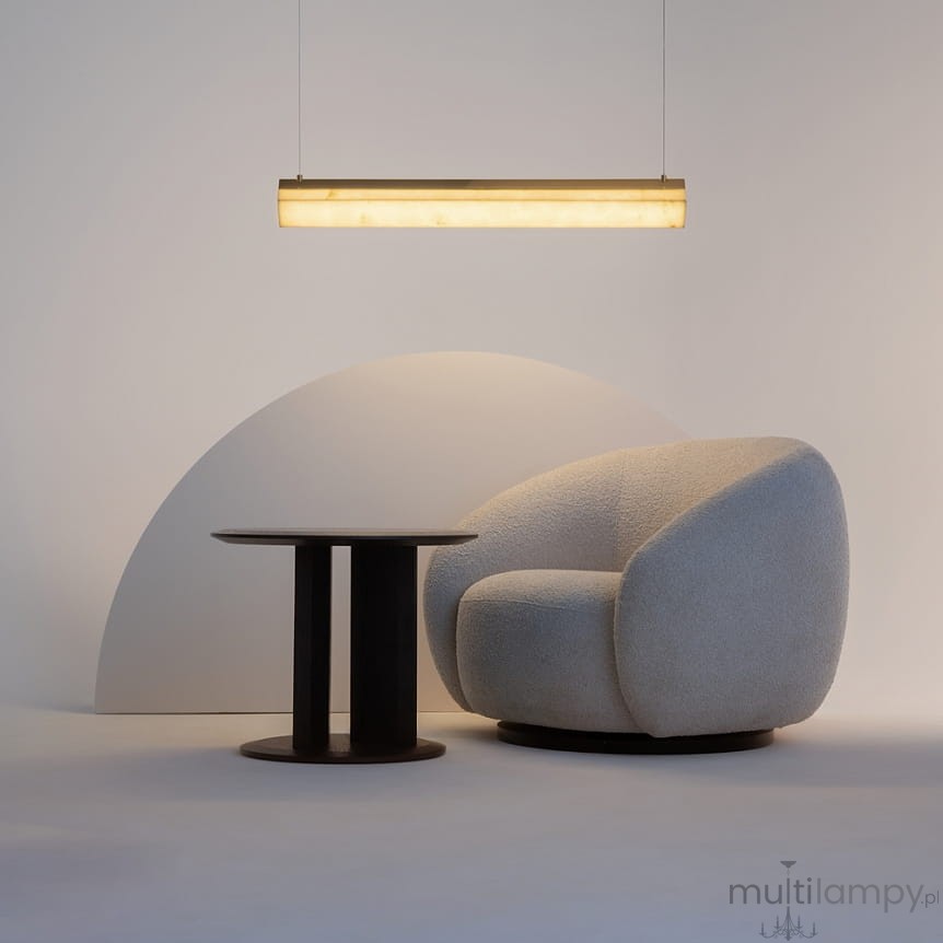 Lampa wisząca LED Petra o mocy 35W i barwie 3000K, w mosiężnej oprawie, zawieszona nad okrągłym stołem w ciemnym kolorze i wygodnym, jasnym fotelu. Tło w neutralnych odcieniach, tworzy nowoczesną aranżację.