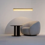 Lampa wisząca LED Petra o mocy 35W i barwie 3000K, w mosiężnej oprawie, zawieszona nad okrągłym stołem w ciemnym kolorze i wygodnym, jasnym fotelu. Tło w neutralnych odcieniach, tworzy nowoczesną aranżację.