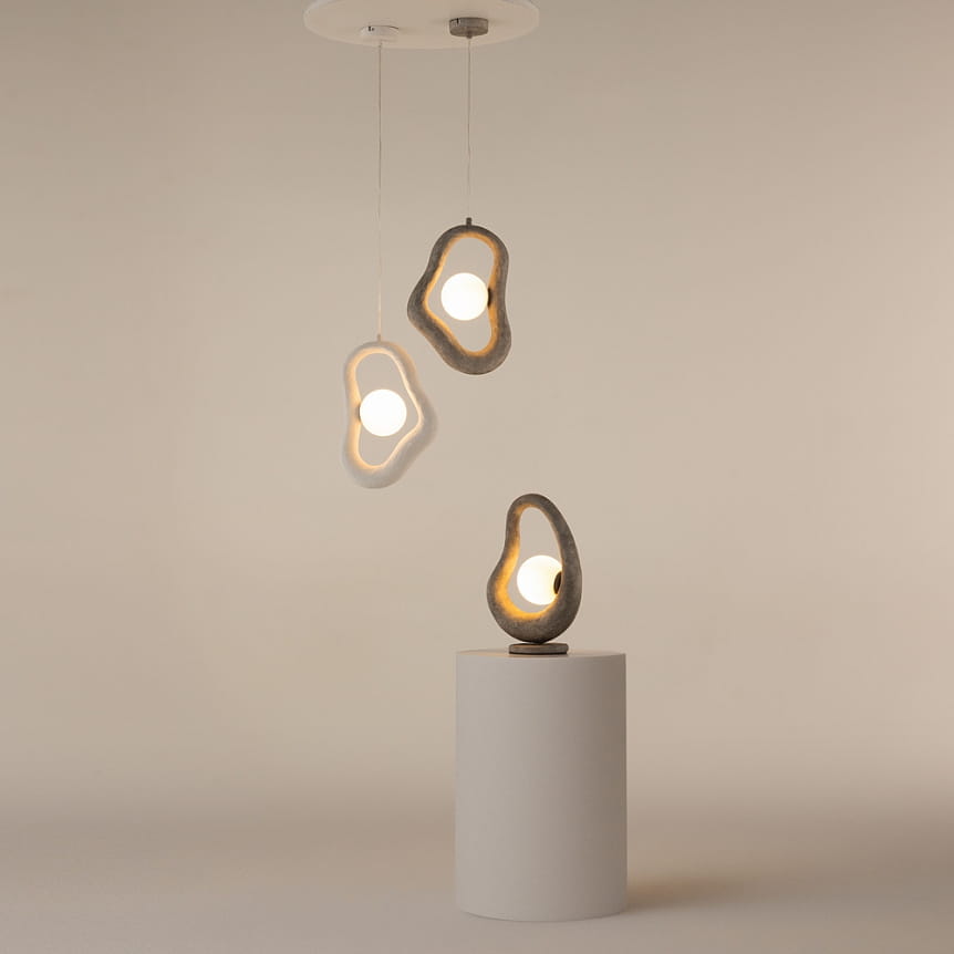 Lampa wisząca Opus w kolorze szarym z jedną oprawą G9. Składa się z trzech asymetrycznych kloszy z betonu, w których umieszczone są żarówki. Dwa klosze wiszą na cienkich linkach, a trzeci stoi na cylindrycznej podstawie.