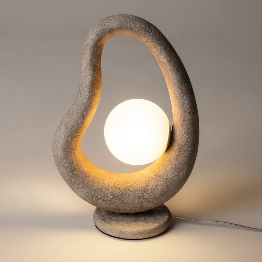 Lampa stołowa Opus MOD380TL-01GR w szarym kolorze z unikalnym, organicznym kształtem. Wewnątrz znajduje się okrągła żarówka G9, emitująca ciepłe światło. Ujęcie frontalne, lampa stoi na okrągłej podstawie, na neutralnym tle.