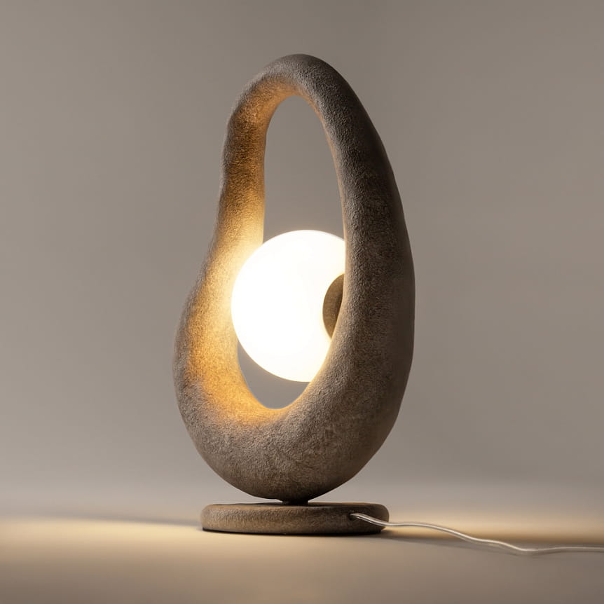 Szara lampa stołowa Opus MOD380TL-01GR z okrągłym kloszem, umieszczona w oprawie o organicznym kształcie. Ujęcie z boku, ukazujące podświetlenie oraz detalowe wykończenie materiału. Kabel zasilający widoczny u podstawy.
