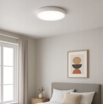Plafon LED Luvo o średnicy 27,5 cm, mocy 18W, barwie światła 4000K, w białej obudowie. Zamontowany na suficie w jasnym wnętrzu, w pobliżu okna i łóżka, z minimalistycznym obrazem na ścianie.