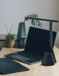 Lampka biurkowa Kiki w kolorze czarnym, z jedną diodą LED 8W, z prostą, nowoczesną konstrukcją. Ujęcie z boku na biurku obok laptopa i dodatków, z widoczną podstawą w kształcie stożka. Tło jasne, minimalistyczne.
