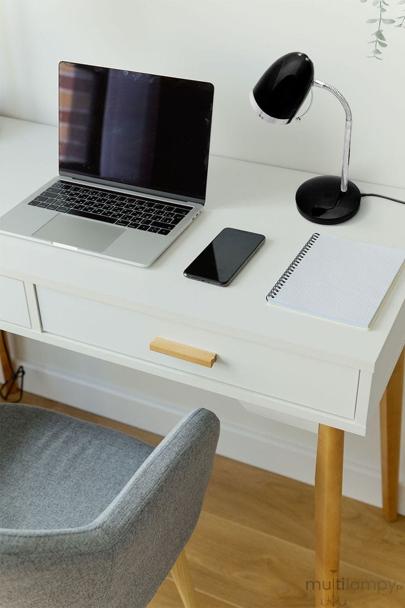 Lampka biurkowa Kajtek, czarna, z metalowym ramieniem, umieszczona obok laptopa na białym biurku. Widoczna na zdjęciu podstawa oraz klosz, z giętkim ramieniem, umożliwiającym regulację kąta. W tle notebook, smartfon i notatnik.