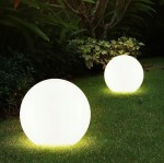 Lampa solarna ogrodowa Ballo D20/H53 LED w kształcie dużej, białej kuli, leżąca na trawie. Dwie lampy emitują miękkie, białe światło, otoczone zielenią roślin. W tle widoczne krzewy, a tło jest naturalne i zróżnicowane.