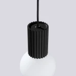 Czarna lampa wisząca Halo z oprawą w kształcie cylindrycznym z rowkami, zawieszona na czarnym przewodzie. Dolna część lampy to biała, okrągła kula. Ujęcie z góry, ukazujące detal mocowania.