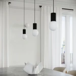 Czarna lampa wisząca Halo z jedną oprawą G9, z trzema kulistymi kloszami w białym kolorze, zawieszona na cienkich czarnych przewodach. Lampa w nowoczesnym stylu, umieszczona nad stołem, w jasnym wnętrzu z zasłonami.