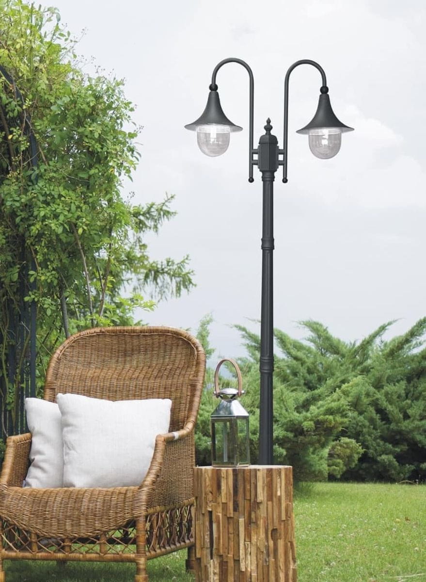 Stojąca lampa ogrodowa Boston H220 w kolorze czarnym, z dwoma kloszami na żarówki E27. Ujęcie z boku, lampy na tle zieleni, w aranżacji z rattanowym fotelem i drewnianą ławą. Widoczne elementy dekoracyjne, jak latarnia obok.