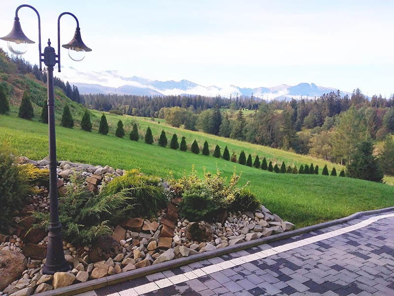 Lampa ogrodowa stojąca Boston H220 w kolorze czarnym, z dwoma źródłami światła E27. Ustawiona na trawniku, w tle widoczna zielona przestrzeń z drzewami i górami. Kąt ujęcia z boku, pokazujący detale lampy i otoczenie.